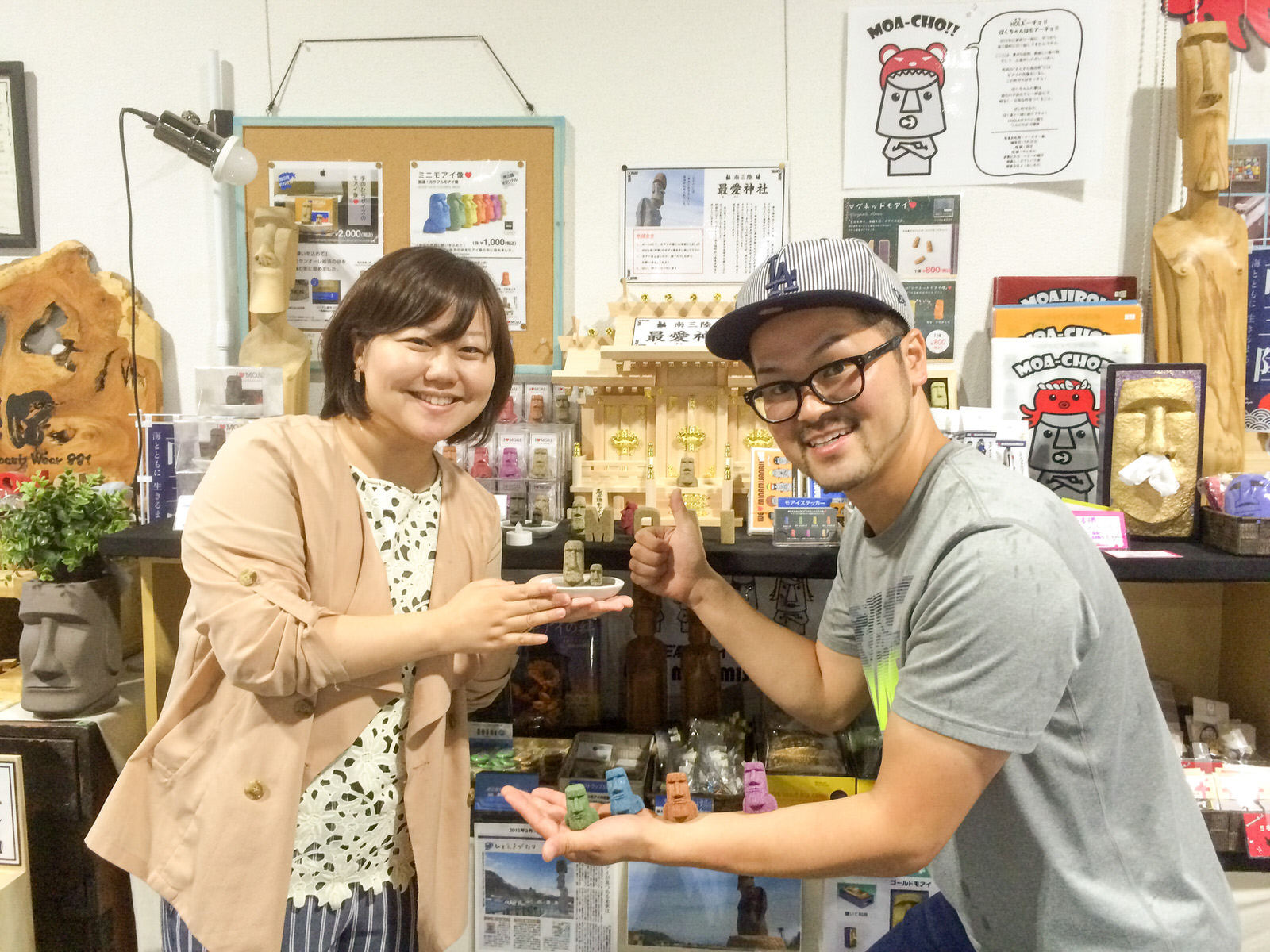 様々なモアイのグッズが並び、神棚が置かれた棚の前で、手にモアイ像が乗った小さな皿を持ったリポーターの女性と、左腕にカラフルな小さいモアイ像を4つ乗せ、右手の親指を立てたキャップをかぶった柳井さんが並んでいる写真