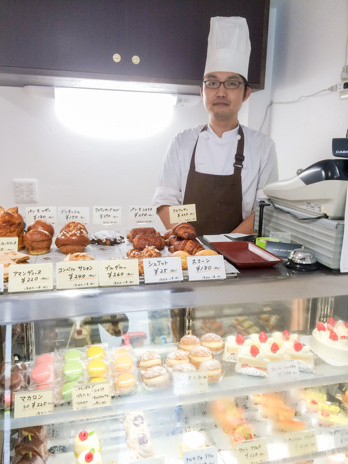 マカロンやシュークリームと様々な種類のケーキが並んだショーケースの上に焼き菓子が並び、その後ろに白いコック服と帽子に茶色のエプロンを着用した三浦さんが立っている写真