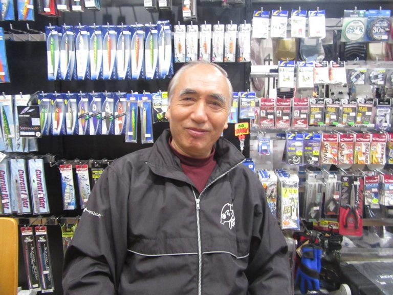 ルアーなどの釣り具が壁一面に陳列された店舗の中で、黒いジャンパーを着た男性が笑顔を見せている様子をアップで撮影した写真