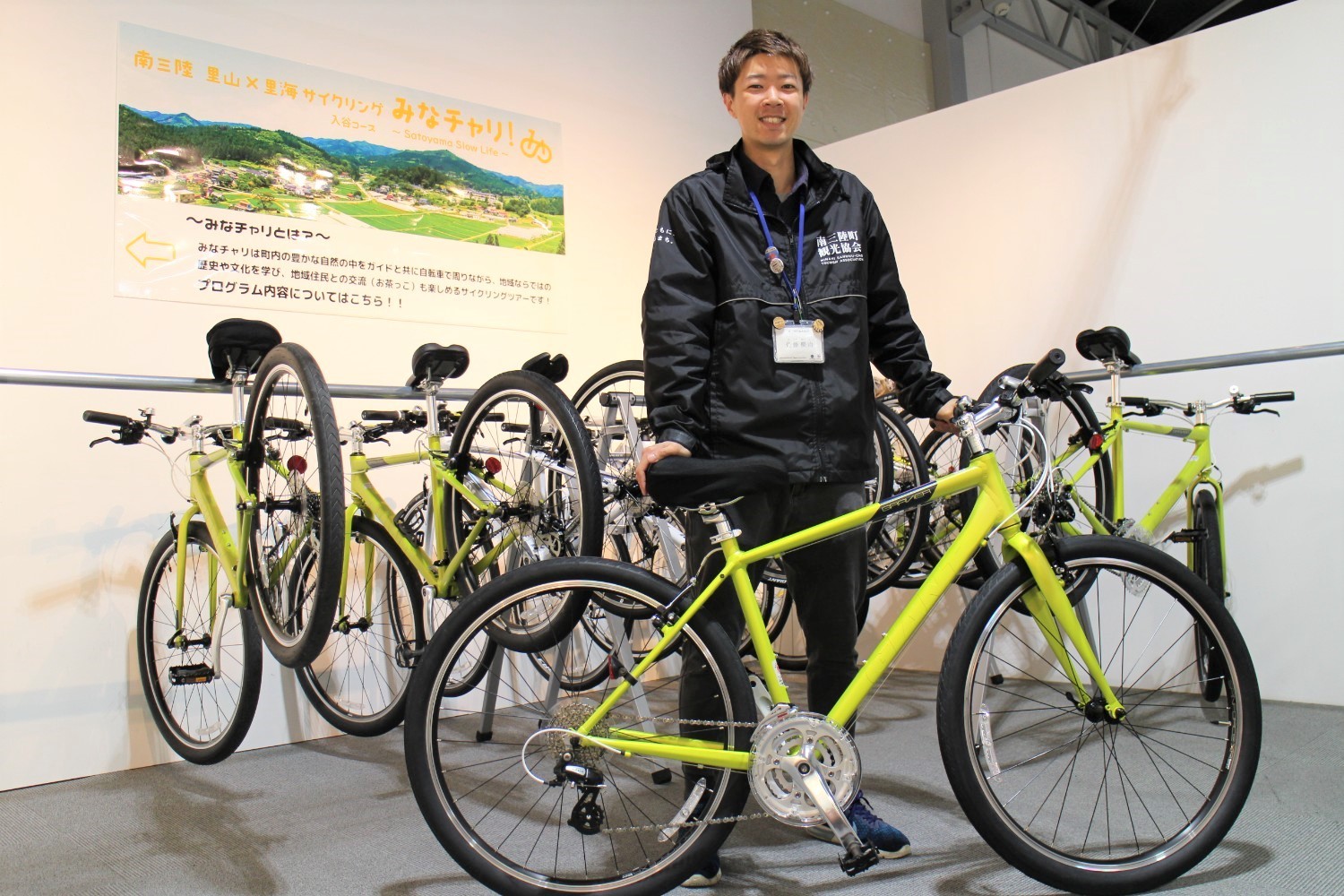 黄色い自転車が何台も展示された室内で、南三陸町観光協会のジャンパーを着た佐藤さんが自転車に手を添えて笑顔で立っている写真