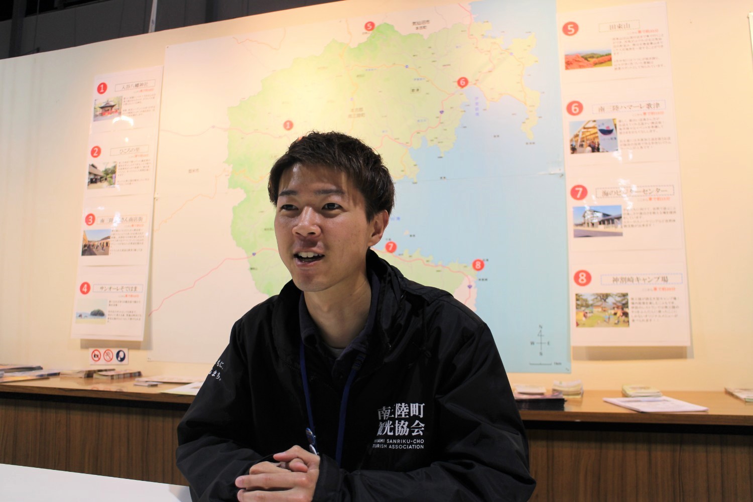 観光スポットが表示された大きな地図が掲示された壁の前に座り、南三陸町観光協会のジャンパーを着た佐藤さんが笑顔で話している写真
