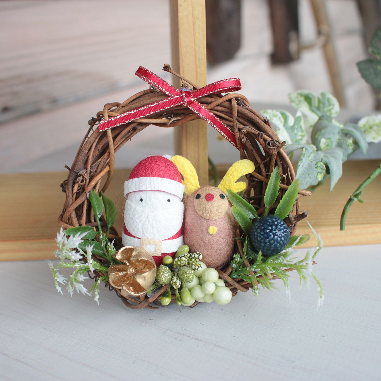 小さなサンタクロースとトナカイのまゆ細工の人形がクリスマスリースの中央に飾られ、周囲に赤いリボンや緑の葉があしらわれている写真