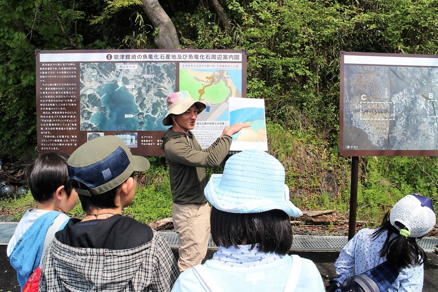 地形図や化石の写真が載った案内板が設置されている前で、帽子をかぶった案内人の男性が子どもたちに地図や説明図を見せながら解説している写真
