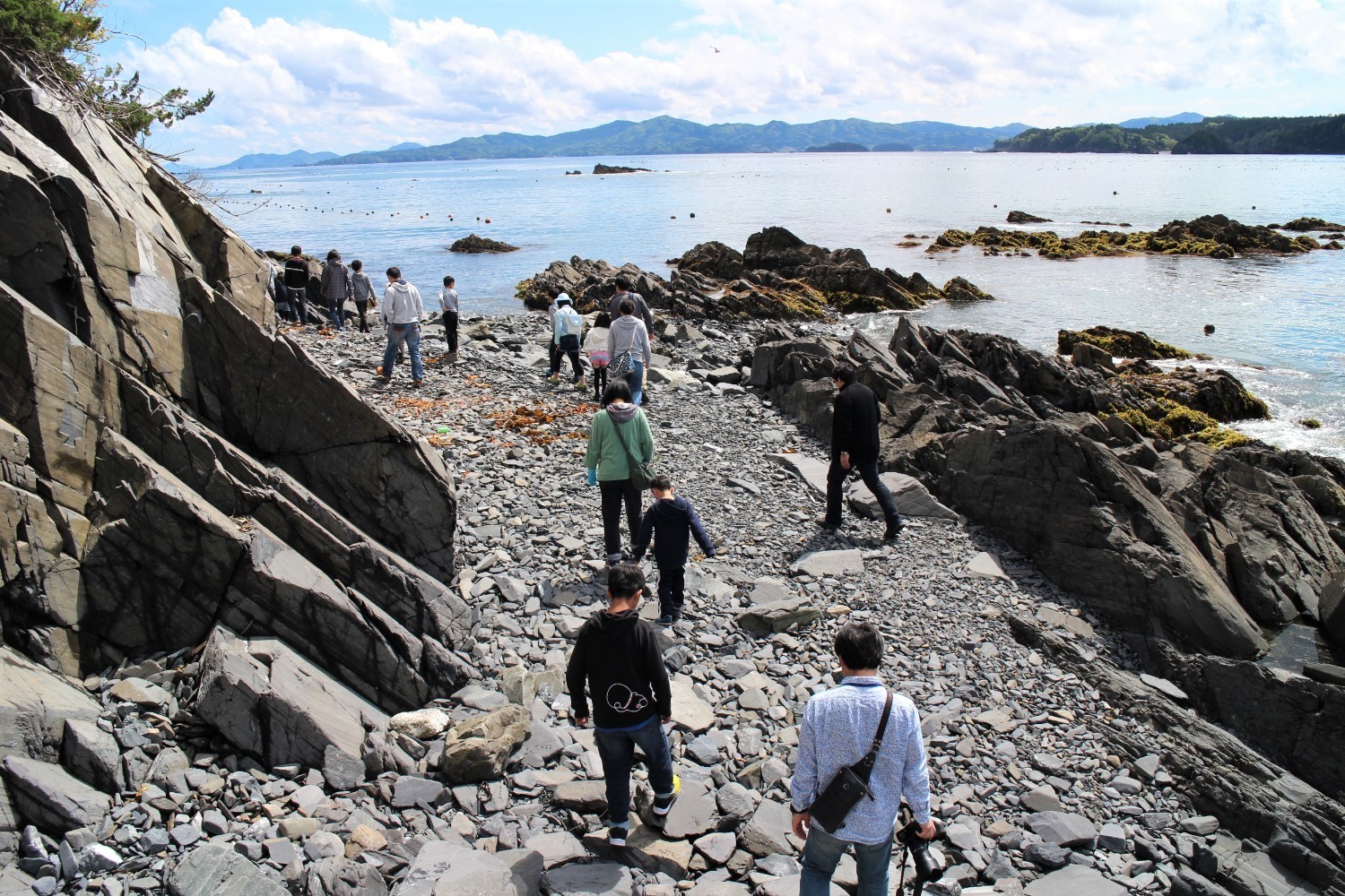 遠くには青い海と島々が広がっている岩場の海岸で、親子の参加者たちが散策して歩いている写真