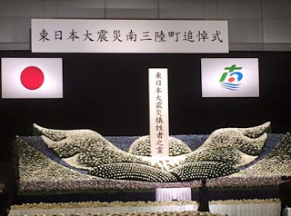 「東日本大震災南三陸町追悼式」の横断幕の下、日本国旗と南三陸町の旗が掲げられたステージいっぱいに白菊の大きな生花装飾が広がり、その中央に「東日本大震災犠牲者之霊」と刻まれた慰霊の碑が立っている写真