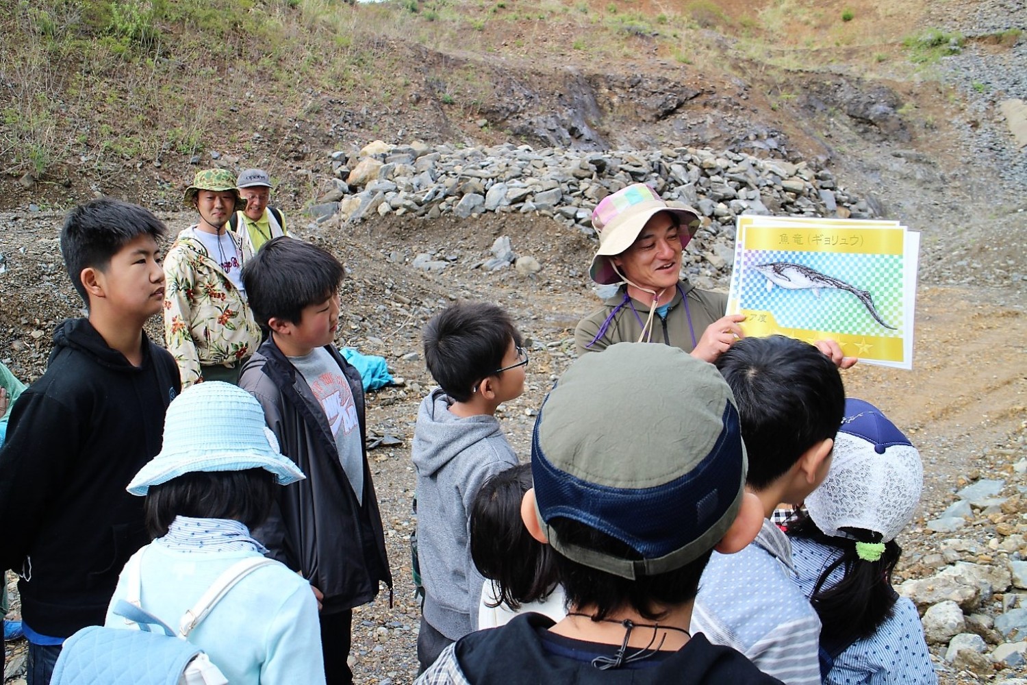 案内人の男性が尾が長くてお腹が膨らんだ魚竜のイラストが描かれたボードを手に説明しており、集まった子どもたちが興味深そうに話を聞いている写真