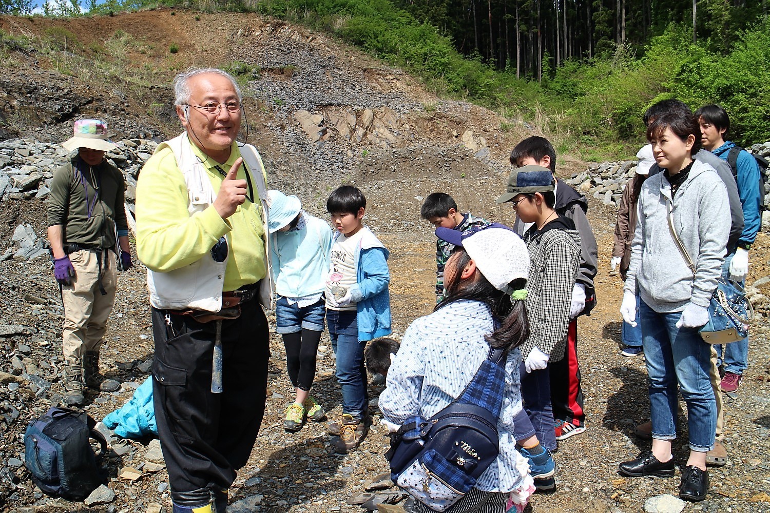 屋外の石が多く転がる場所で、牧口さんが子どもたちに向けて人差し指を立てながら説明をしており、手袋をつけた子どもや大人たちの参加者が話を真剣に聞いている様子の写真