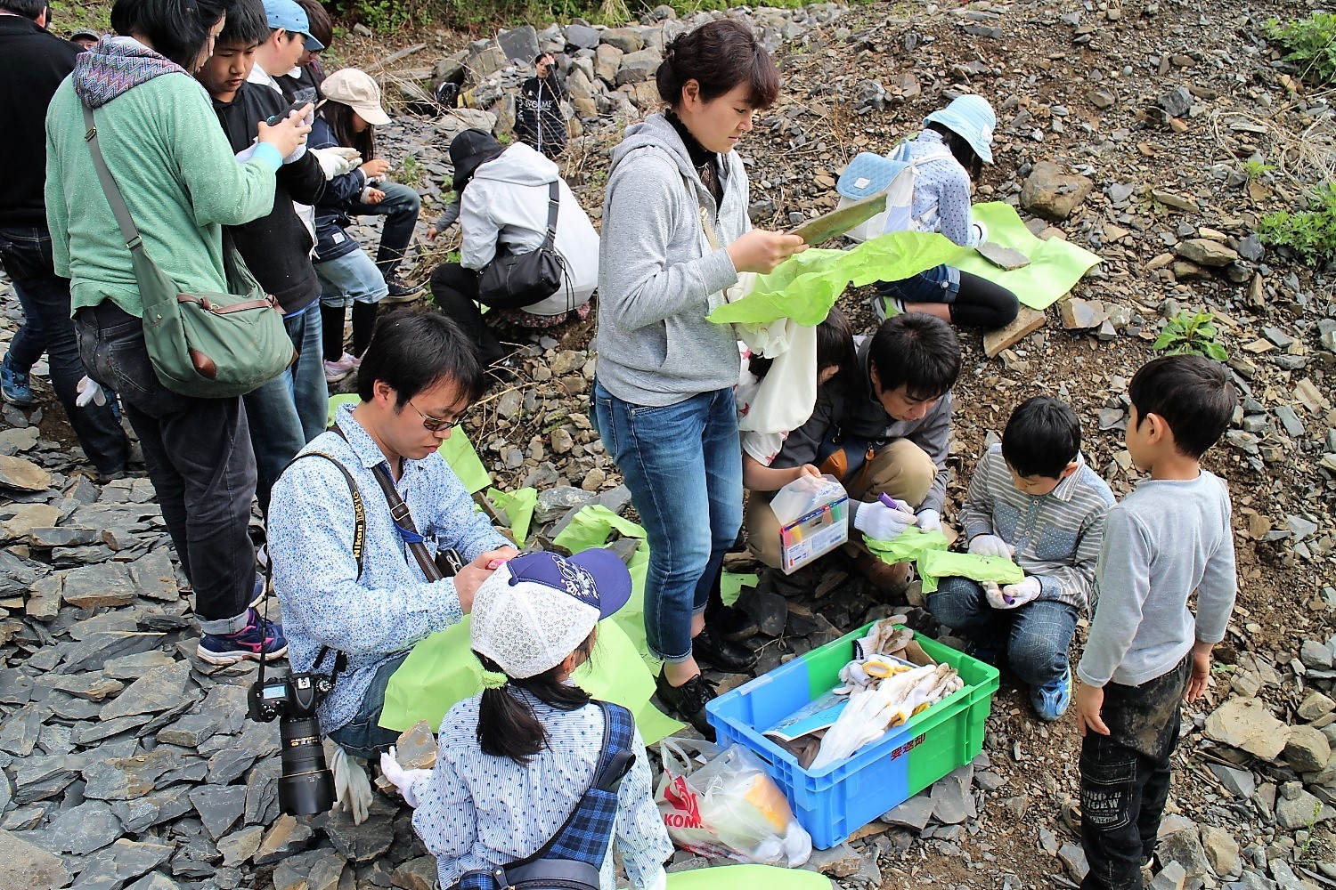 緑色の包装紙や軍手が入っている道具箱が置かれた屋外で、多くの親子連れが地面に座ったりしゃがんだりしながら石を包む準備をしている様子の写真
