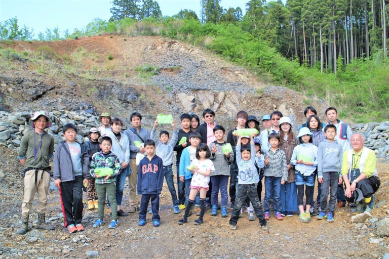 山のふもとの岩場で、何人かが緑色の布にくるまれた化石を掲げている、子どもから大人までの約30人の参加者が並んでいる集合写真