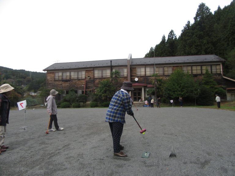 木造の古い校舎を背景に、広場で高齢者たちがグラウンドゴルフを楽しんでいる様子を捉えた写真