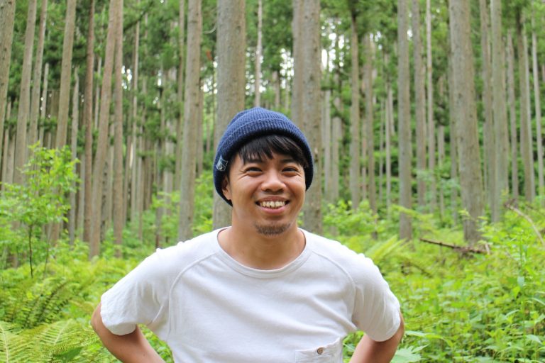 白いTシャツに青いニット帽をかぶった笑顔の平山太一さんが、緑の森林の中に立っている様子をアップで撮影した写真