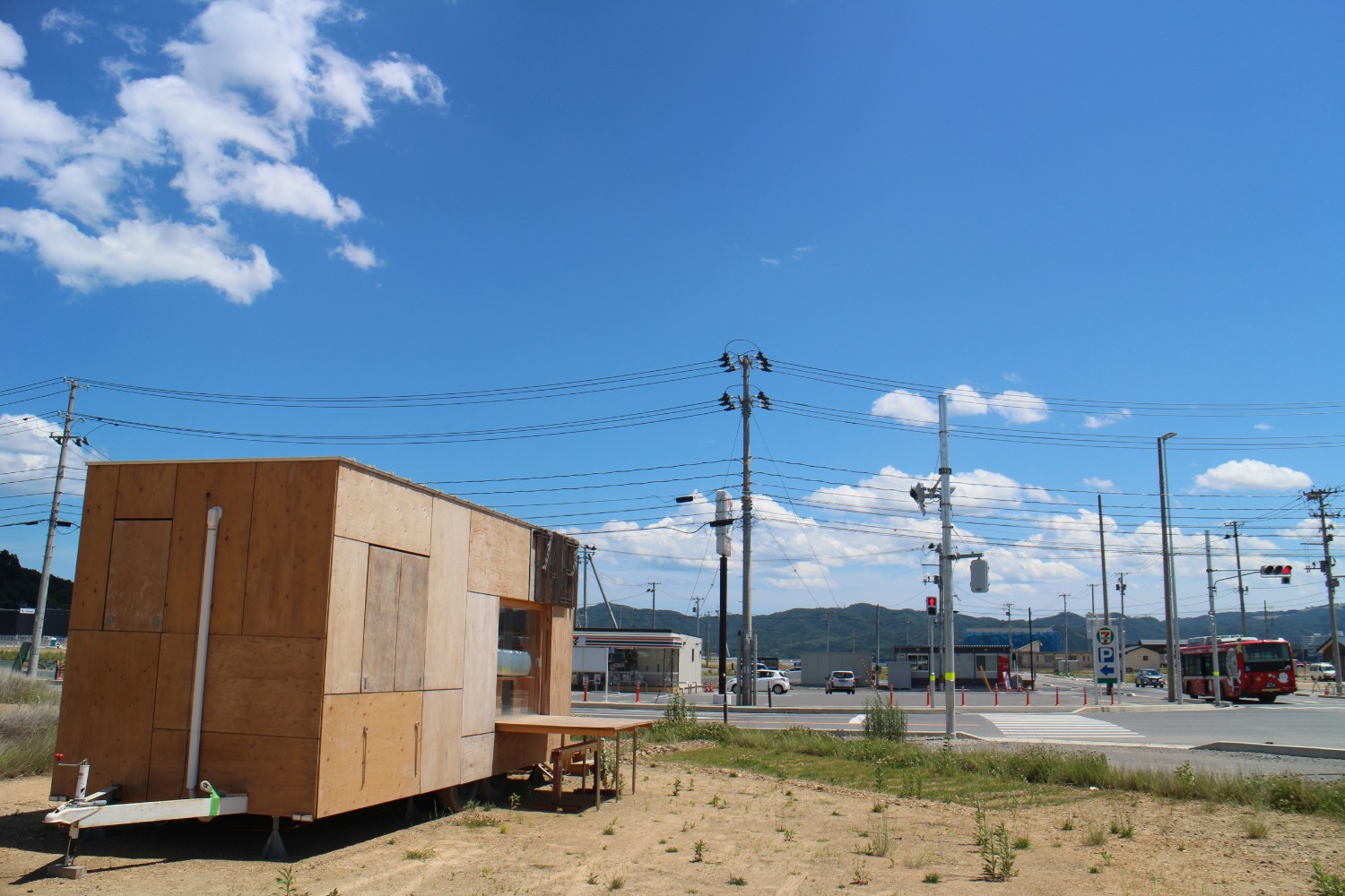 信号機やバス、遠くの山々と青空が見えている道路沿いの空き地に、木製の小さなトレーラーハウスが設置されている写真