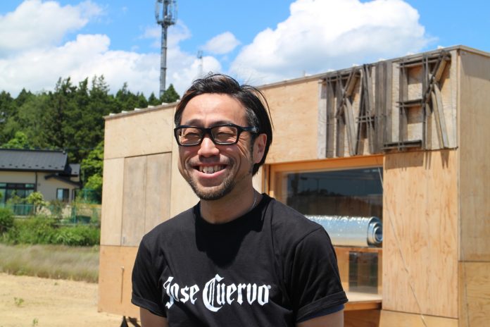 青空が広がっている木製のトレーラーハウスの前で、黒いTシャツを着た縁の眼鏡をかけた短髪の井原健児さんが屋外で笑顔を見せている様子をアップで撮影した写真