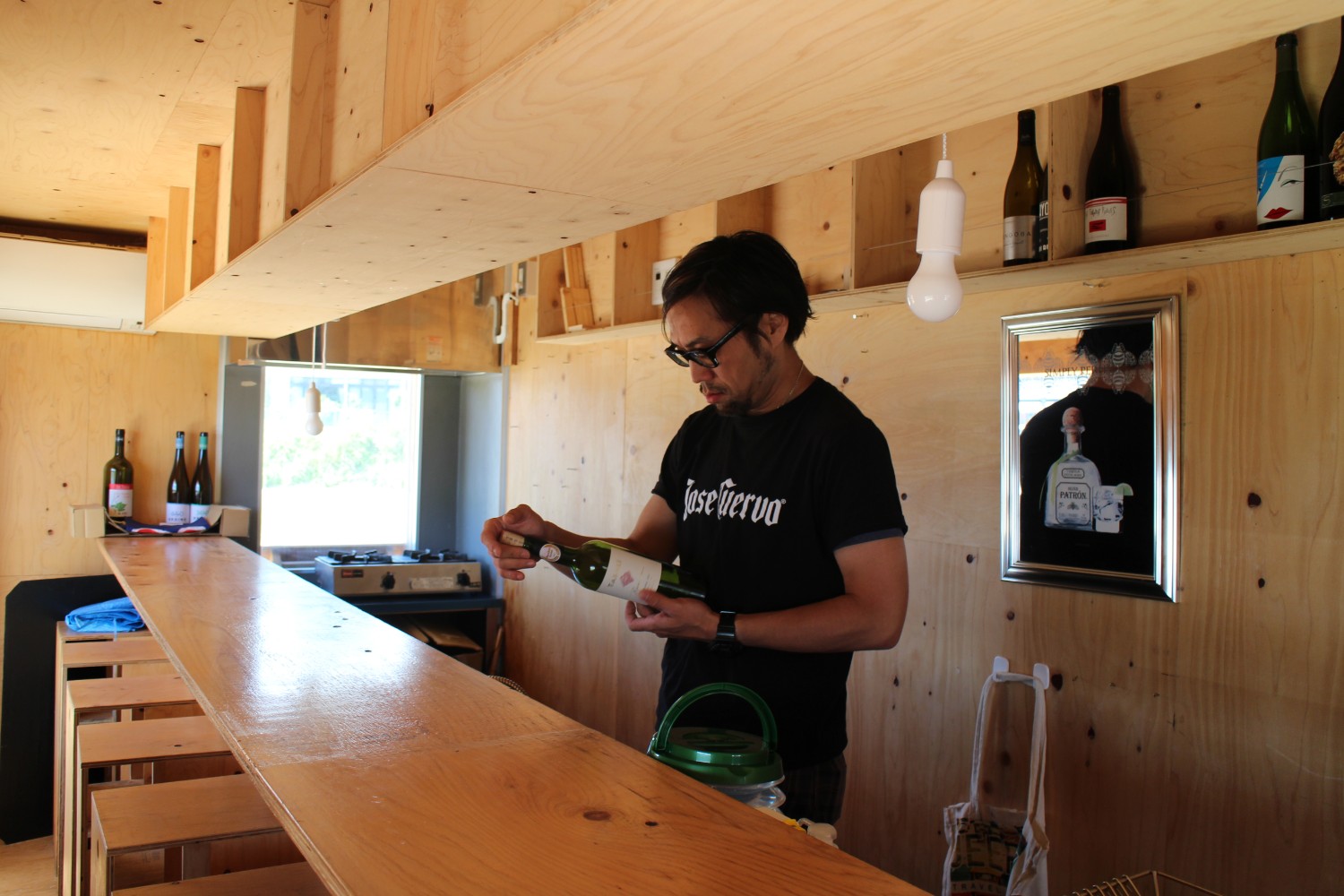 棚や窓、ワインボトルが並んだ簡素な内装の木のカウンターのある小さな店内で、黒いTシャツを着た井原健児さんがワインボトルのラベルを見ている写真