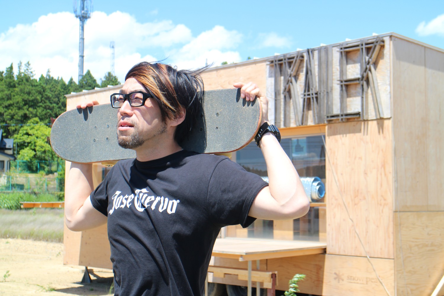 緑の木々が広がっている木製のトレーラーハウスの前で、黒いTシャツの井原健児さんがスケートボードを両肩に担ぎながら青空を見上げている写真