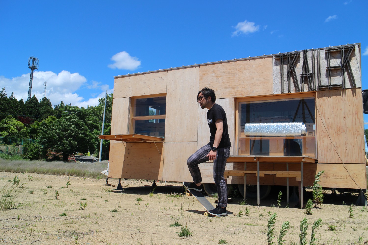 背景には森と空が広がっている木製のトレーラーハウスの前で、黒いTシャツの井原健児さんがスケートボードに足をかけて乗ろうとしている写真