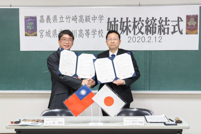 「嘉義県立竹崎高級中学 宮城県立志津川高等学校 姉妹校締結式」と書かれた幕が貼られた黒板の前で、2人のスーツ姿の男性が協定書を開いて持ち、片手で握手を交わしている記念写真