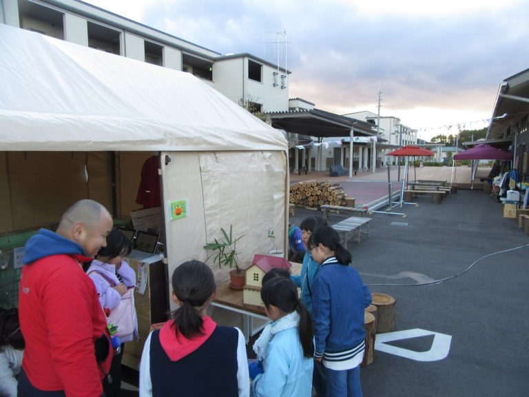 木材が積まれた屋外スペースが広がり、テントの外に置かれた小さな家の模型と植物の前で、男性が5人の女の子たちと話をしている写真