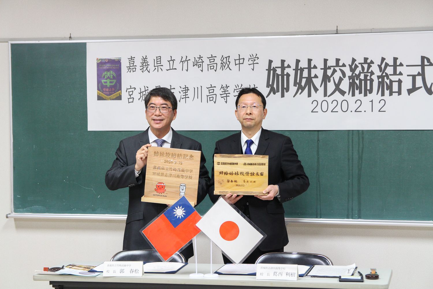 男性2人がそれぞれ木製の記念プレートを持って並んで立っている写真