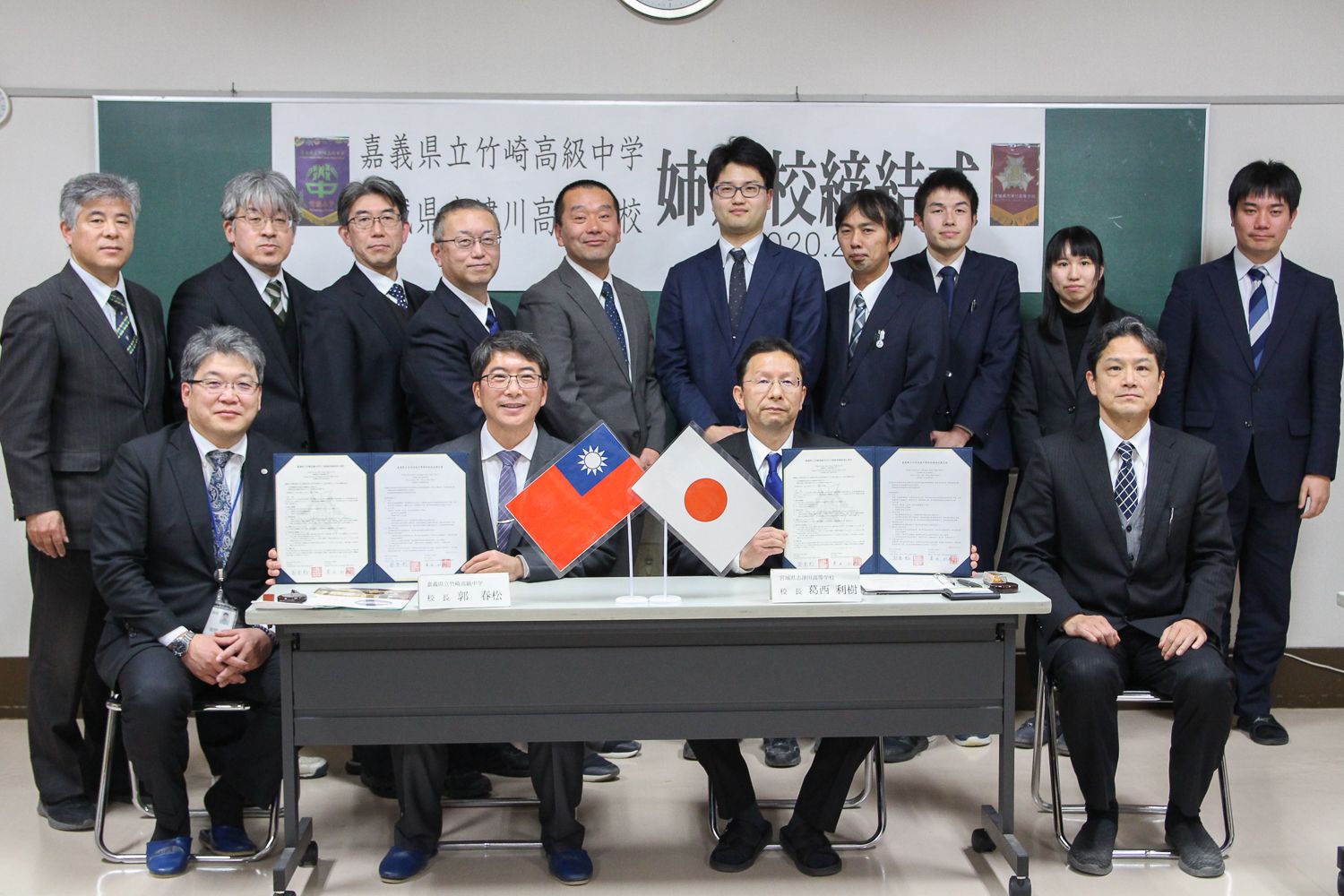 台湾と日本の国旗を前にし、姉妹校締結の書類を掲げて関係者たちが並んで記念撮影している写真