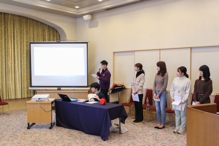 会議室で1人の男性がマイクで説明しており、関係者がプロジェクターを使ってプレゼンテーションをしている学生達の写真