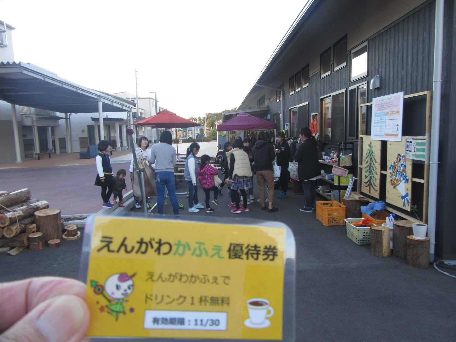 建物の外で多くの人々が集まり、テーブルや屋台が並ぶにぎやかな雰囲気の中、手前で「えんがわかふぇ優待券」を手元に映した写真