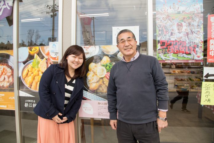 食べ物のポスターなどが貼られた店の入り口前で、リポーターの女性と山内正文さんが笑顔で並んで立っている写真