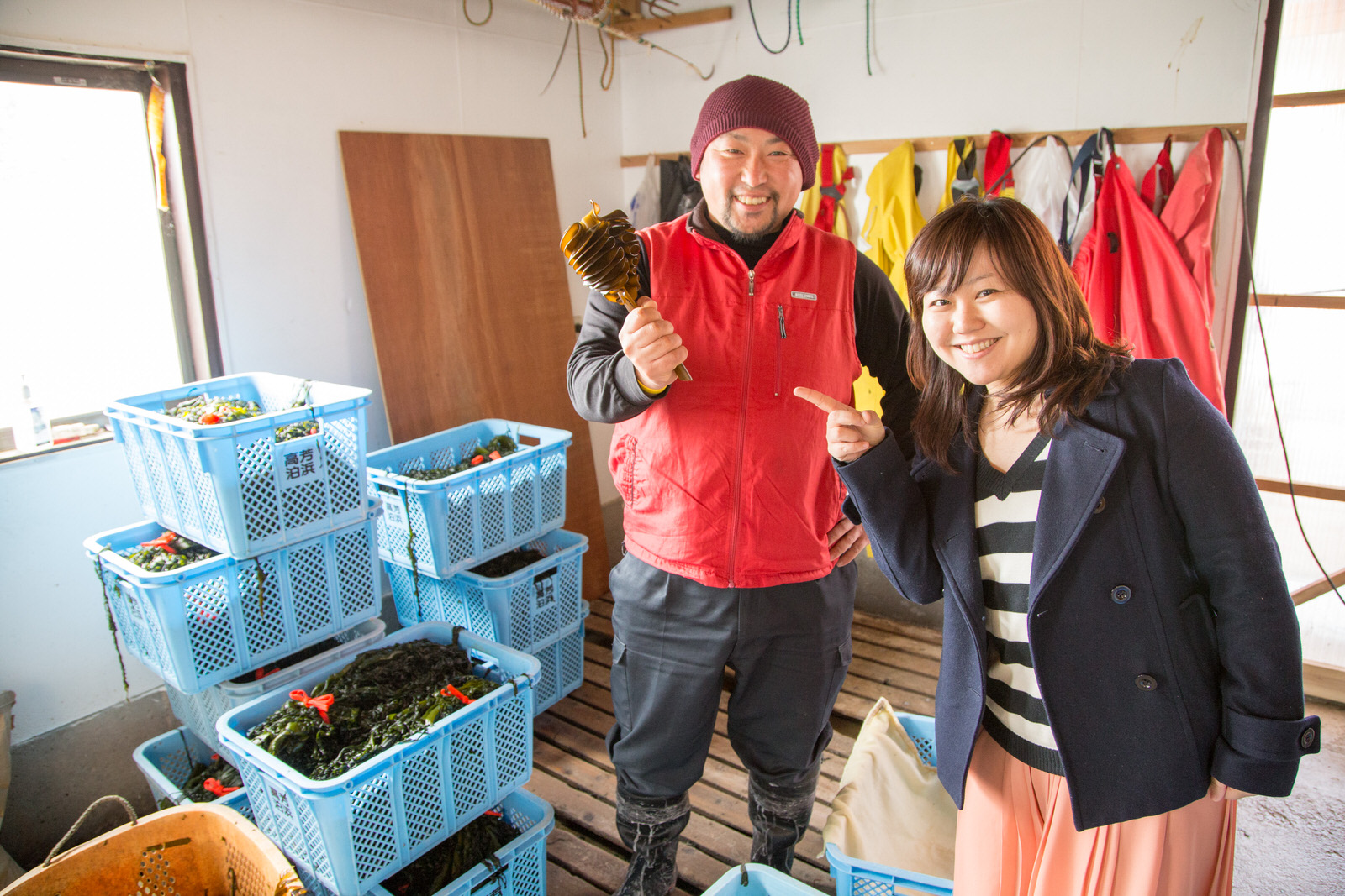 わかめが入った水色のかごがたくさん積まれた室内で高橋さんとリポーターの女性が笑顔で並んでいる写真