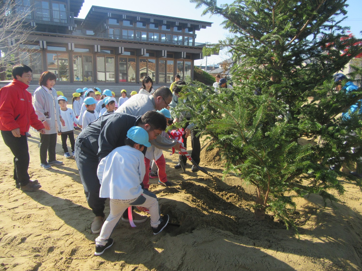 大きな建物を背景に、大人と子供たちが屋外の土の地面でスコップを使い協力して木を植えている様子の写真