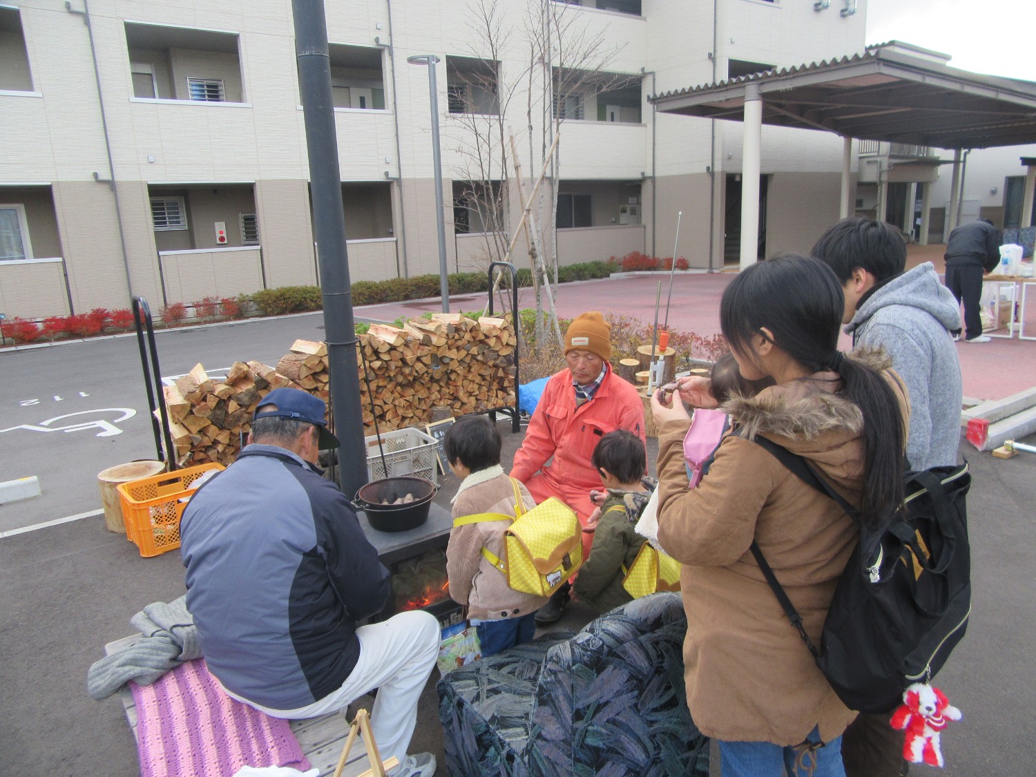 マンションの駐車場で、数人が焚き火を囲んで暖を取っており、背景には積み上げられた薪の山と建物が見える様子が写っている写真