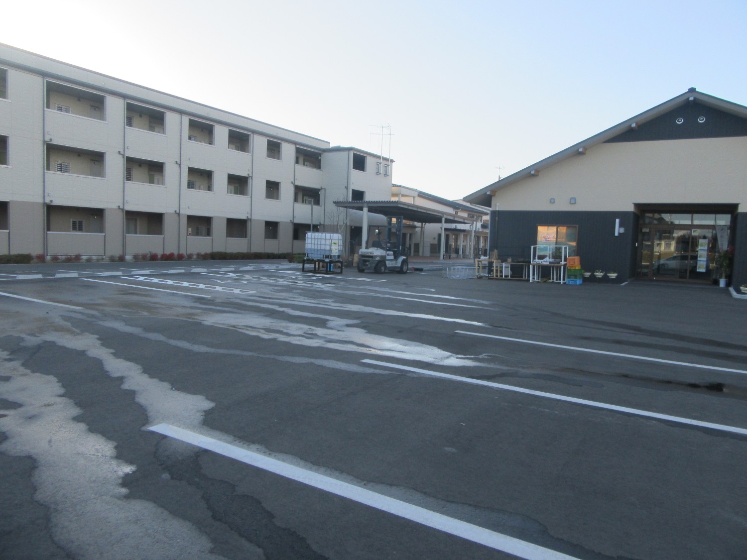 広い駐車場の右奥に三角屋根の「めぐるステーション」の外観が写り、左側には3階建ての建物がある写真