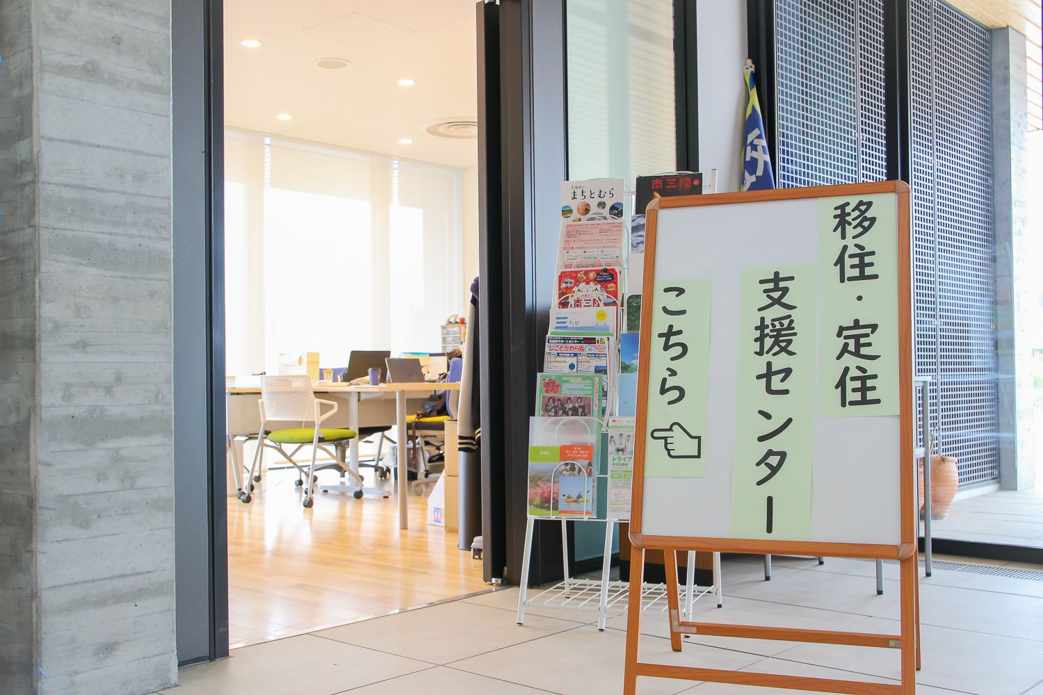 屋内の明るいオフィスの入口付近に、「移住・定住支援センターこちら」と書かれた白い掲示板が置かれている写真