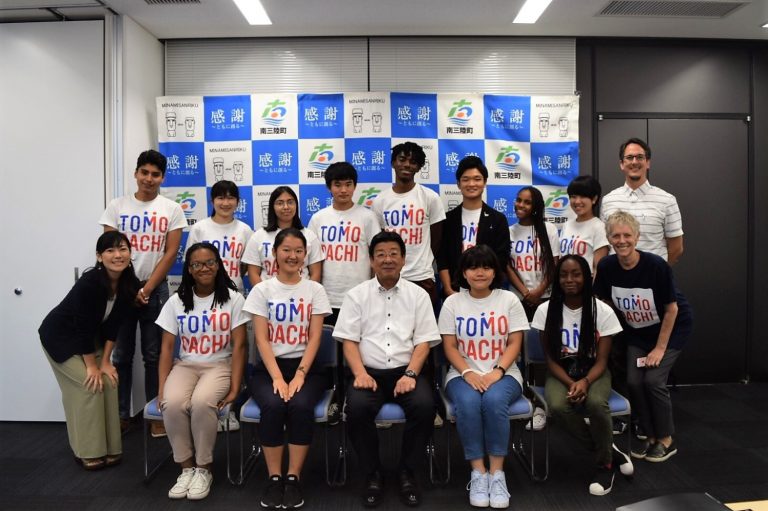 南三陸町のバックパネルの前で、青や赤色で「TOMODACHI」と書かれたTシャツを着た多国籍の若者たちが、佐藤町長を中央に2列に並んでいる記念写真