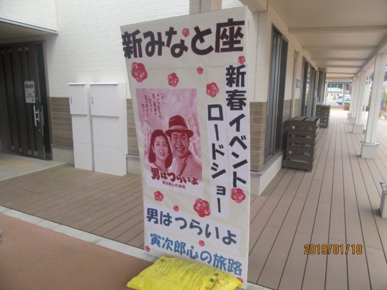 建物の軒下の通路の角に、映画『男はつらいよ』のポスターを掲げた立て看板が置かれている様子を写した写真