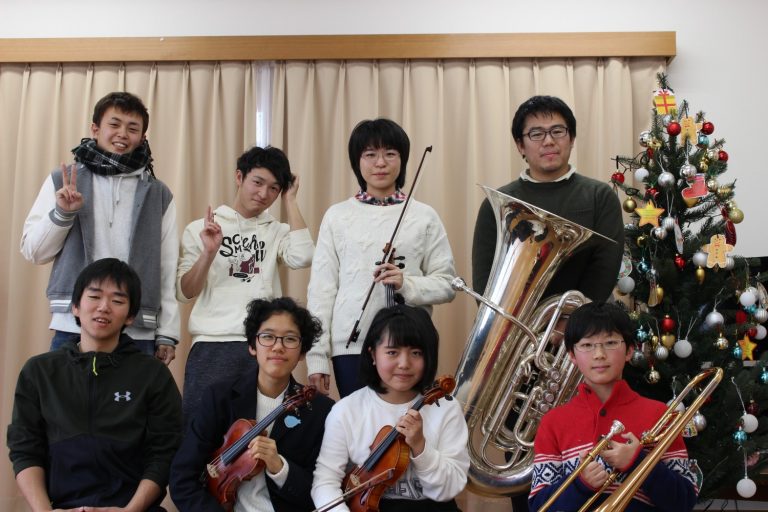 クリスマスツリーの横で、東北ユースオーケストラの男女8人が楽器を持って笑顔で写っている集合写真