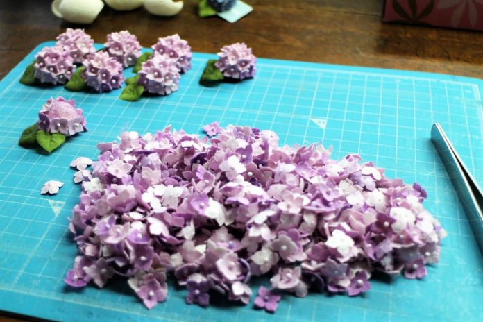 作業マットの上に小さな紫色のアジサイの花びらが大量に並べられ、奥にはそれらを組み合わせて作られた立体的なアジサイの花と葉のパーツがいくつも置かれている、繊細な手作りクラフトの制作途中を写した写真