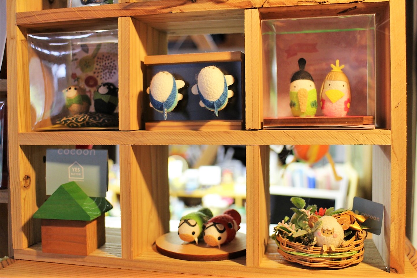 木製の六つの仕切りがある飾り棚に、フェルトや布で作られた小さな鳥の人形や家のミニチュアなどが並べられている写真