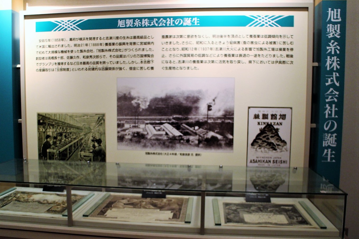 旭製糸株式会社の誕生について解説した展示パネルで、パネル上部に日本語の詳細な説明文があり、中央に煙を上げる工場のモノクロ写真と工場内部で働く人々の写真、右側に「ASAHIKAN SEISHI」と記された絹織物のラベル風の画像が配置されているほか、パネルの前にはガラスケースに収められた複数の古い資料や写真が並んでいる展示室の様子を写した写真