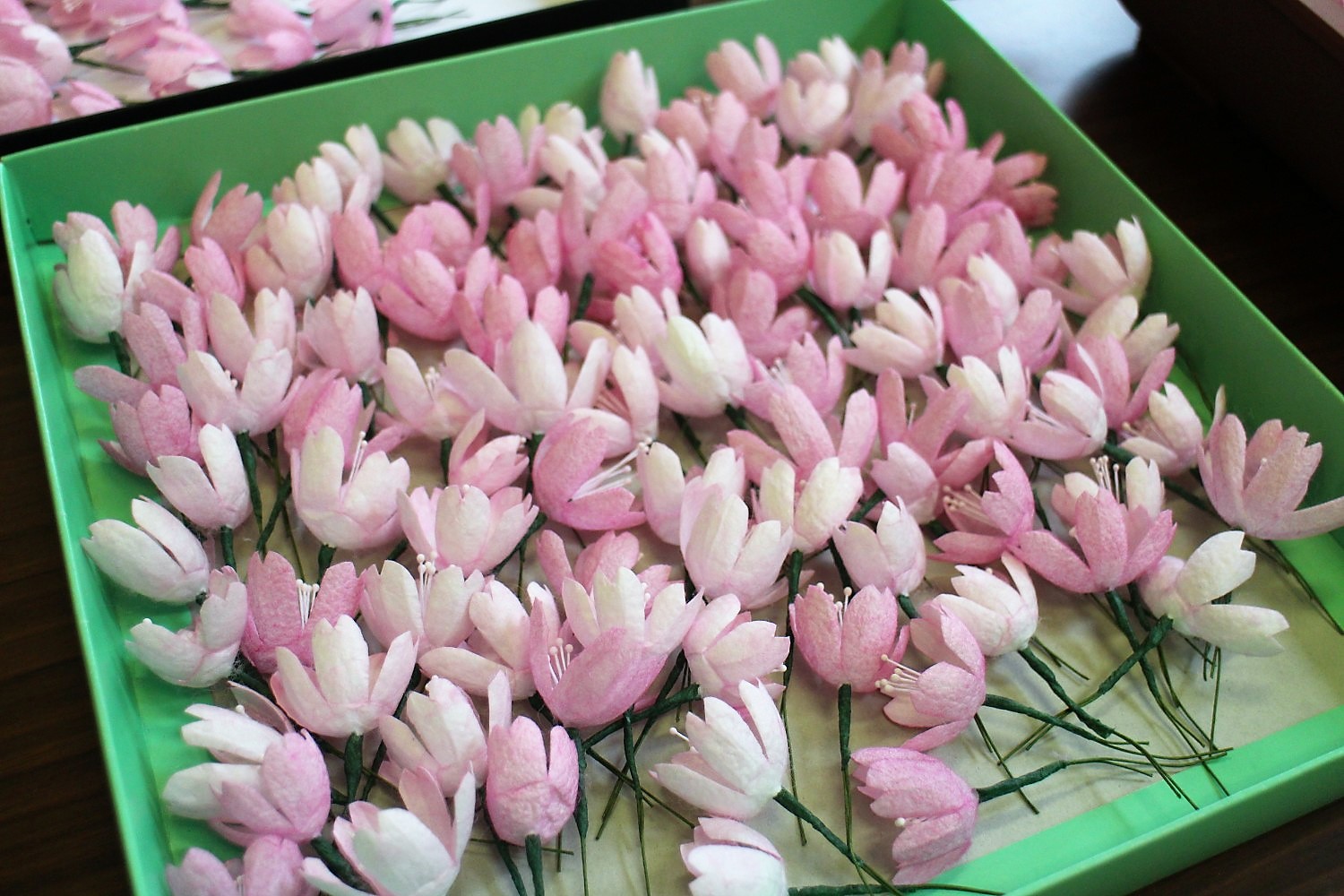 淡いピンク色から白色にかけての小さな花の形をした繊細な造花が緑色の箱の中にたくさん並べられている写真