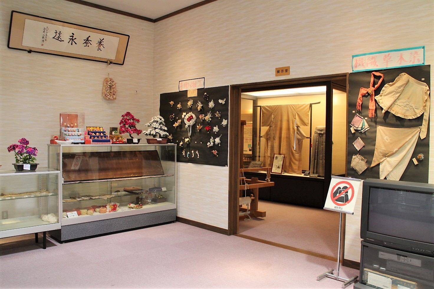 薄いベージュ色の壁とカーペット敷きの床を持つ室内で、左側にはガラスケースの中に手芸品や造花が展示され、中央には展示室の入口がありその奥に着物や織物が飾られ、右手前にはテレビとビデオデッキのセットが置かれているが、入口付近に撮影禁止を示す赤い斜線のマークが掲示されている写真