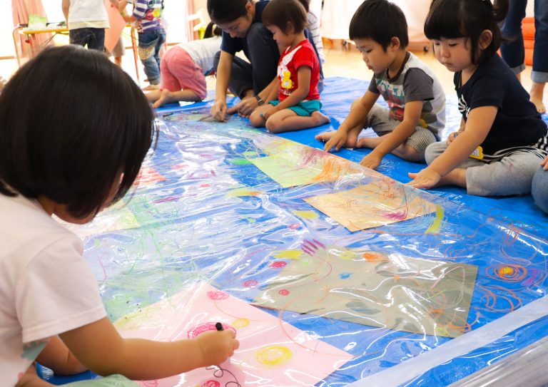 青いビニールシートの上に透明なシートが広げられ、周りに座ってお絵描きを楽しんでいる子供達の写真