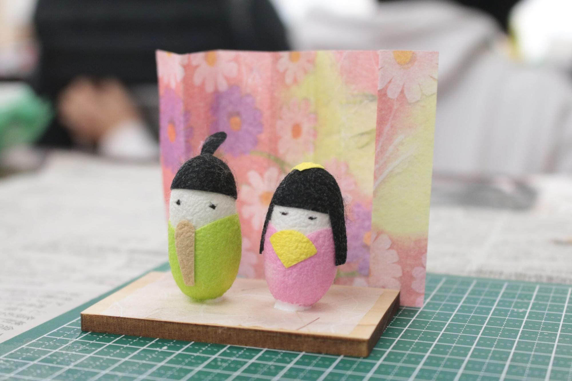 緑とピンクの胴体をした小さな和装のまゆ細工のひな人形が二体、和紙の花柄の背景の前に木の台座の上に飾られている写真