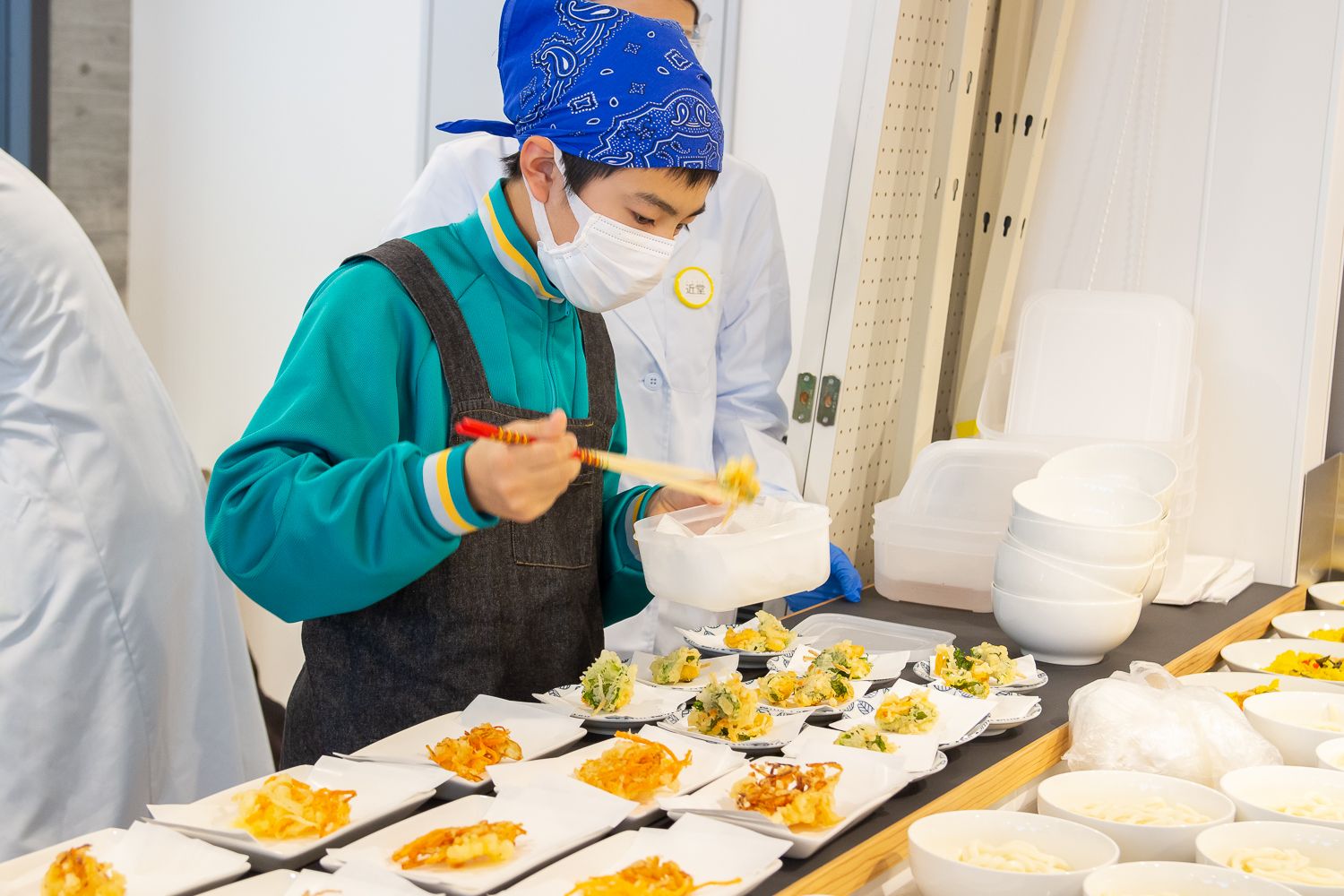 青いバンダナとマスクをつけた子どもが、エプロン姿で調理台の上に並べた白い皿に野菜の天ぷらを盛り付けている写真