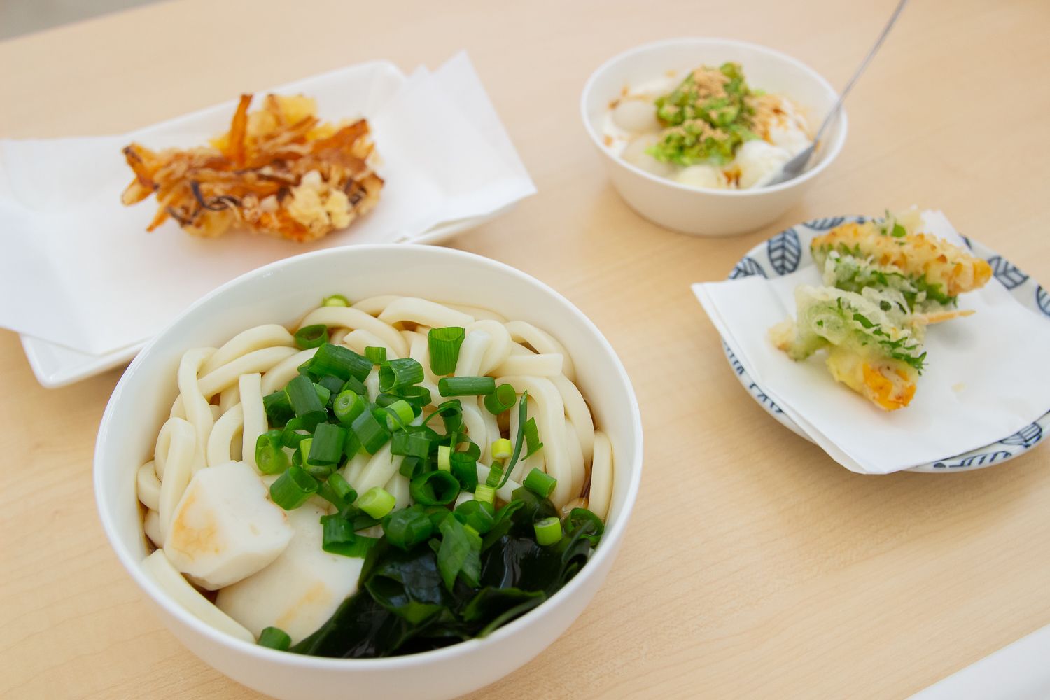 テーブルの上に、刻んだネギがのった三陸わかめうどん、ホヤと大葉の天ぷら、地元食材のかき揚げ、スプーンが添えられた手作りずんだアイスがそれぞれ白い器や皿に盛り付けられている料理の写真