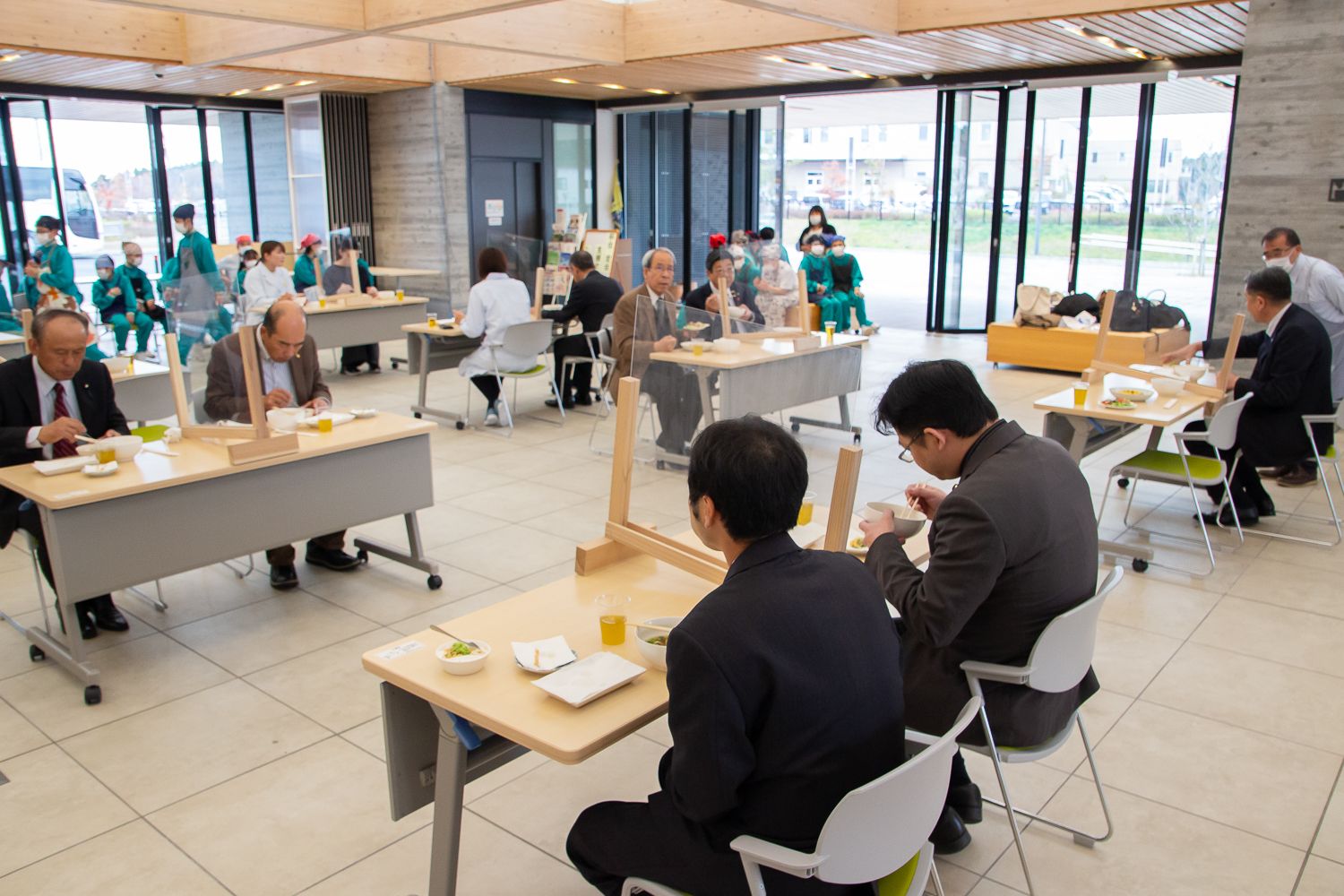 広い室内で、透明のパーテーションが間に設置されたテーブルに座ったスーツ姿の大人たちが食事をしており、奥にはエプロン姿の子どもたちが座って見守っている写真