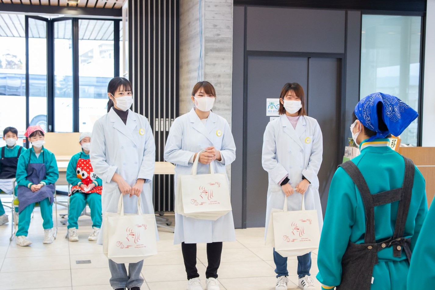 後ろの椅子にエプロン姿の子どもたちが座って見ているなか、エコバッグを持った白衣を着た三人の女子学生が生徒代表の子どもと向かい合って立っている写真