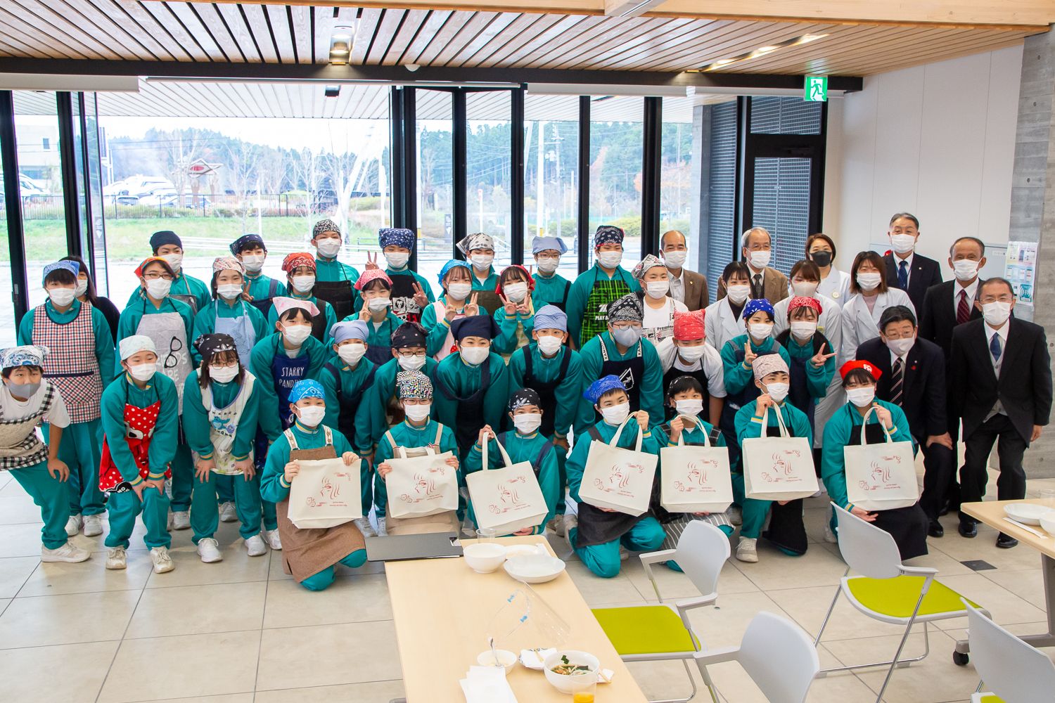 前列に座った7人の子どもたちはエコバッグを手に持ち、エプロンを着た生徒たちと白衣やスーツ姿の大人たちがマスクをつけて大人数で並んでいる集合写真