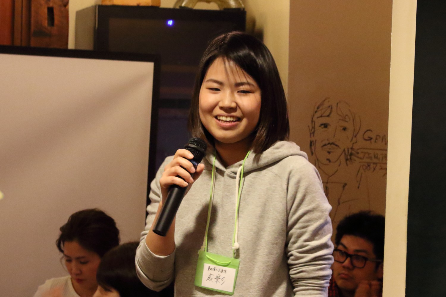 壁に人の顔の絵が描かれ、数人の人が座って聞いているなか、灰色のパーカーを着た女性がマイクを持って笑顔で話している写真