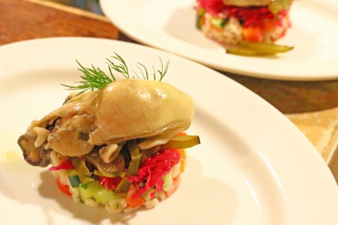 プレートの中央に細かくカットされたカラフルな野菜と穀物を土台にして大きな牡蠣が乗っており、上にはディルが飾られている料理の写真