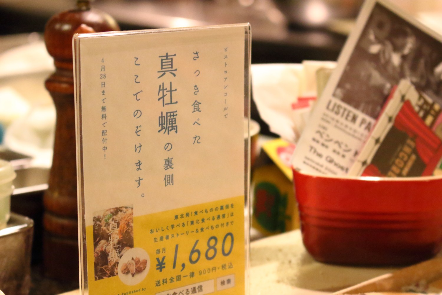 店内のカウンターに立てられた透明の案内板に「ビストロアンコールでさっき食べた真牡蠣の裏側 ここでのぞけます」と書かれ、下に料金や提供情報が記載されている情報誌と食材をセットにした案内の写真